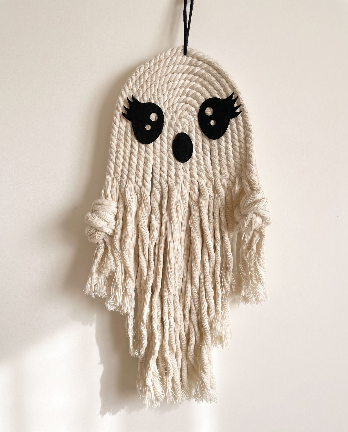 Cream cotton-rope macrame ghost wall hanging with lash eyes and long fringe on an off-white wall